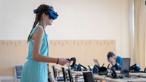VR 기반 수업에 참여하여 VR 헤드셋을 착용하고 한 손에는 컨트롤러를 들고 책상 앞에 서 있는 학생