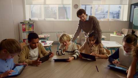 각자 태블릿이 있는 어린 학생 6명이 교실의 공용 테이블에 앉아 있고, 교사가 한 학생의 어깨 너머로 진행 상황을 바라보고 있음