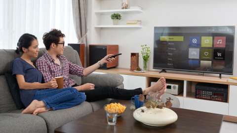 두 사람이 거실의 소파에 나란히 앉아 있습니다. 한 사람이 TV 리모컨을 사용하여 평면 TV에 표시된 스트리밍 서비스 선택 메뉴를 탐색하고 있습니다